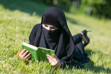 Islamic woman reads a book on a lawn in a park. Muslim student girl preparing for an exam reads a...