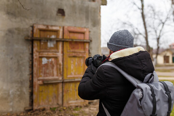 Man in warm jacket and cap with back pack standing back to camera and taking photographs of abandoned building outdoors.