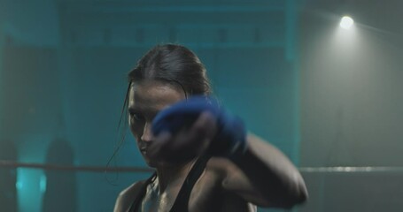 Close up of young attractive Caucasian woman boxer punching with fists to camera and striking on dark ring. Portrait of female fighter boxing and looking concentrated straight. Kickboxing concept. - Powered by Adobe
