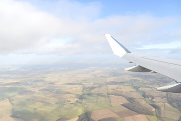 High angle view of landscrape of green land under white airplane wing with clouds in blue sky. View from airplane window