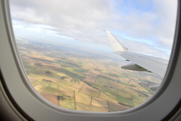Landscape of green land under white airplane wing with clouds in blue sky, looking Through window of airplane.
