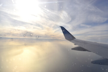 Airplane wing above clouds with sun setting over horizon. Looking through aircraft window.