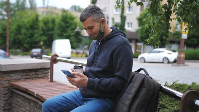 A man with a medical mask pulled down on his chin is sitting on a bench in the city, using a smartphone and drinking coffee from a paper Cup.