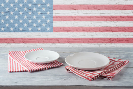 Empty Plate With Stripes Tablecloth On Wooden Table Over Brick Wall With American Flag. 4th Of July USA Independence Day Mock Up For Design.