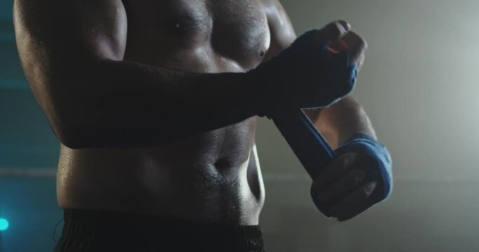 Close up of Caucasian strong male kickboxer putting on straps on his hands before fighting on ring. Man boxer getting ready to fight ir training in dark. Kickboxing concept. Preparation to box.
