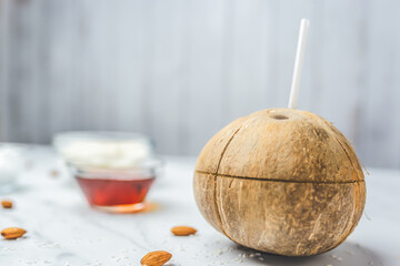 Close-up of a whole vibrant round coconut with a drinking straw and a marked cut for cracking on the table with food preparing ingredients: syrup, butter, and desiccated coconut in the background.