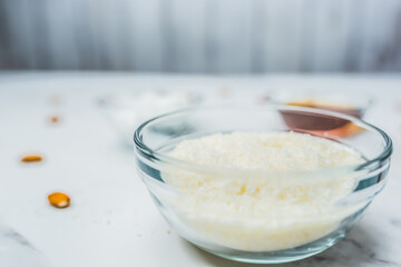 Close-up of a bowl with desiccated/dried coconut in focus with ingredients in a glass bowl on the table: maple syrup, coconut butter, and almond nuts. Concept of the homemade chocolate preparation.
