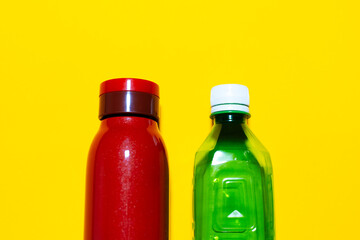 Close-up of two closed bottles. Reusable steel thermo water bottle of red color and disposable plastic bottle of green. Studio background of yellow.