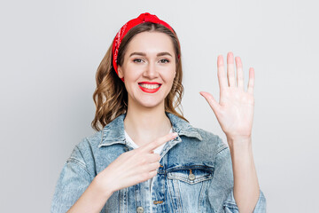 Student girl in denim jacket, bandana smiles and points to her left hand isolated on gray...