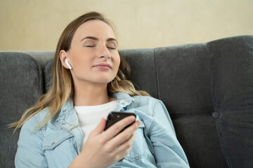 Young woman sitting listening to music with earphones on her mobile phone at home as she relaxes with closed eyes on a sofa.
