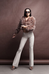 Young elegant girl posing in fluffy pink cape with artificial fur and beige flare trousers on brown background