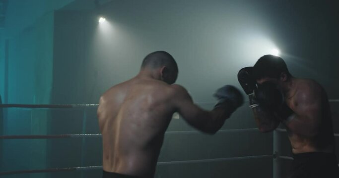 Two Caucasian professional young shirtless male kickboxers fighting at dark boxing ring. Sportsmen boxers kickboxing in darkness at training. Fight sport concept.