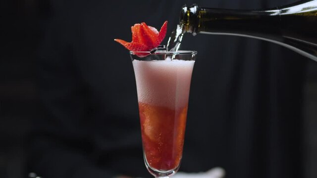 Making Strawberry Rossini Cocktail. Close up bartenders hands pouring champagne into a glass decorated with strawberry flower on a skewer. The foam rises in a glass