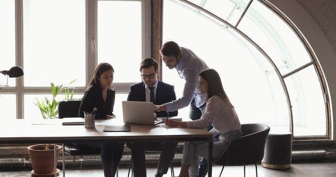 Serious business team of four multiethnic coworkers discuss digital market strategy using laptop at office meeting table. Diverse employees group talking doing online research data analysis together.