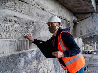 A male engineer in a construction helmet and antiviral mask inspects an emergency building for restoration or repair.