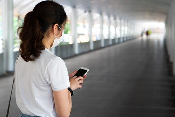 young asian woman wearing face mask using smartphone and walking in the city during covid-19 or coronavirus outbreak. social distancing and new normal lifestyle concept