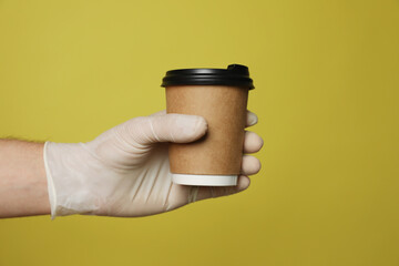 Courier in protective gloves holding cup with drink on yellow background, closeup of hand. Delivery service during coronavirus quarantine