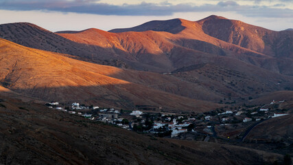 Letztes Sonnenlicht fällt auf eine hügelige Wüstenlandschaft mit Bergen, Tälern und einer Siedlung im Tal