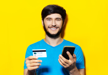 Studio portrait of young smiling guy in blue shirt, looking at camera, using smartphone and credit card on background of yellow color.