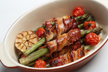 Oven baked asparagus wrapped with bacon in ceramic dish on white table, closeup