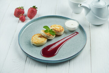 Homemade cheesecakes or Syrniki with jam, fresh fruits and sour cream on a blue plate on white wooden background. Ukrainian cuisine. Breakfast food