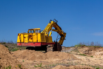 Obraz premium Yellow excavator during earthmoving at open pit on blue sky background. Construction machinery and earth-moving heavy equipment for excavation, loading, lifting and hauling of cargo on job sites