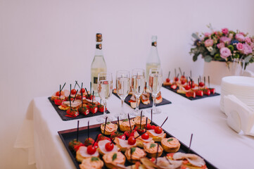 Four glasses with champagne, black square trays with canapes. Reception, catering, clean dishes, napkins, two bottles of champagne. The table is covered with a white tablecloth. Bouquet of pink flower