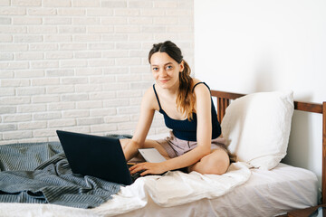 A young woman, using a laptop, communicates over the Internet with a client in her bed, while the virus is quarantined.Cozy office workplace,Remote work, e-learning concept.freelancer