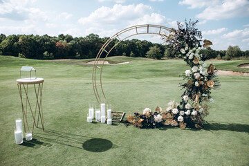 A wedding arch stands on a bare field and is decorated with tropical plants, golden flowers and...