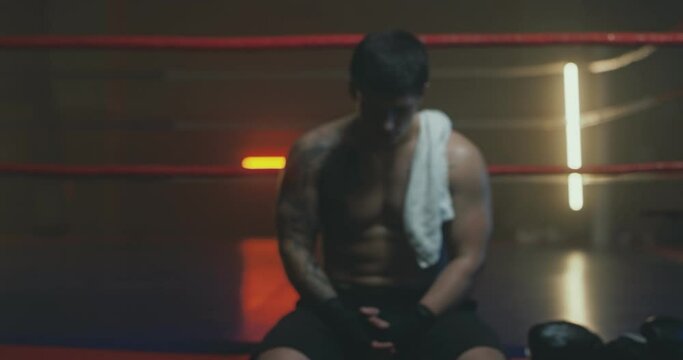 Camera zooming in on young Caucasian male shirtless sweaty sportsman sitting at ring and being upset of loosing fight. Lost in box concept. Boxer is dissapointed. Blurred. Slow motion.