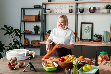 Happy blond middle aged female looking in smartphone camera while cooking