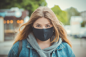 Closeup portrait of a girl in a medical mask. Quarantine of a pandemic coronavirus. Covid 19. Student in a protective mask. Young girl with long hair in a black protective mask in the city.