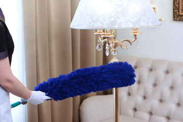 The maid dusts the floor lamp with a large brush. An unrecognizable photo . Concept of cleaning in...