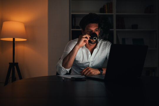 Young businessman drinking coffe and using a laptop computer at his desk in the office or home late into the night