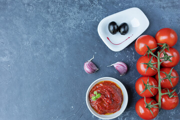 Tomato sauce-ketchup in a bowl with spices, herbs and fresh cherry tomatoes on a branch, on a dark graphite background. The view from the top.