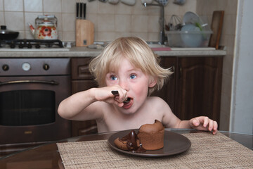 .little boy with blond hair sits at a table in the kitchen and eats with a spoon chocolate fondant cupcake