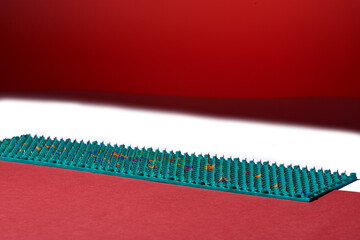Massager in the form of green carpet with metal spikes, needles, lies on red and white backgrounds, diagonal. Abstraction. For relaxation health