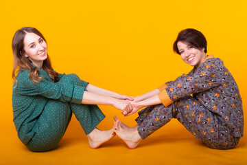 Portrait of two beautiful young girls wearing colorful pyjamas having fun during sleepover isolated over yellow background. Pajama party and hen-party concept