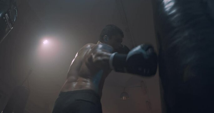 View from below on brutal shirtless young Caucasian strong male boxer with tattoo punching bag in gym at ring and training. Man boxing and exercising concept.