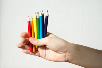 The hand of a white child on a white background holds seven colored pencils. Rainbow color. Drawing - what to do with your child at home.