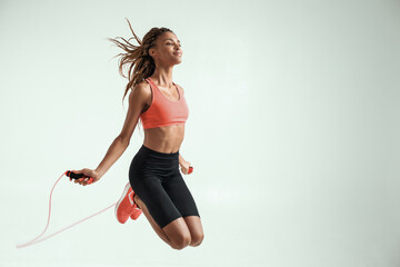 Flying. Happy and young african woman with perfect body skipping rope while exercising in studio...