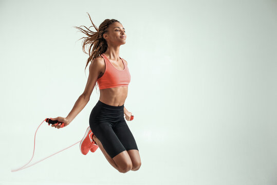 Flying. Happy and young african woman with perfect body skipping rope while exercising in studio against grey background
