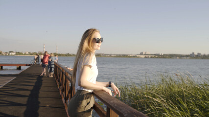 Happy smiling young blond woman in trendy sunglasses looking at summer sunset on river. Cute face model smiling on cityscape lake background. Girl looks joy at camera. Blond hair fly fluttering wind.