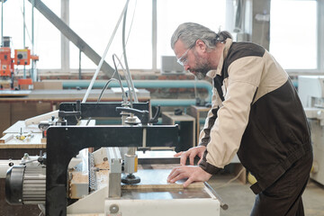 Mature engineer of furniture factory bending over industrial machine in workshop