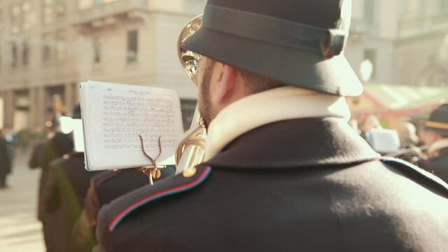 Musician Looks At Sheet Of Paper With Notes And Plays Wind Instrument, Tuba. Back View. Dressed In Police Or Military Uniform, Walking Through The City With A Brass Band. Bright Sunny Day