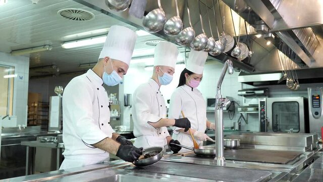 Chefs in protective masks and gloves prepare food in the kitchen of a restaurant or hotel