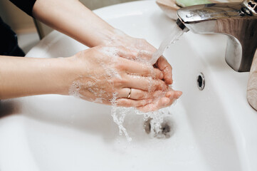 Female hands with soapy foam in a stream of tap water in the bathroom close-up. The concept of...
