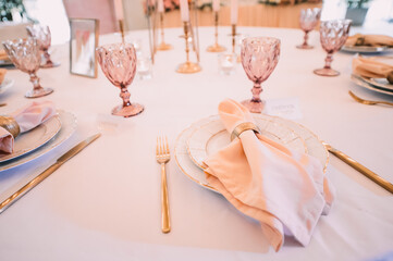Banquet, restaurant. Table setting. White plates with a gold rim, golden cutlery, glasses. Peach-colored napkin with a golden ring. Tall tapers in gold candle holders
