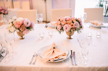 Banquet, restaurant. Table setting. White plates with a gold rim, golden cutlery, glasses. Peach-colored napkin with a golden ring. Peach pink floral arrangements