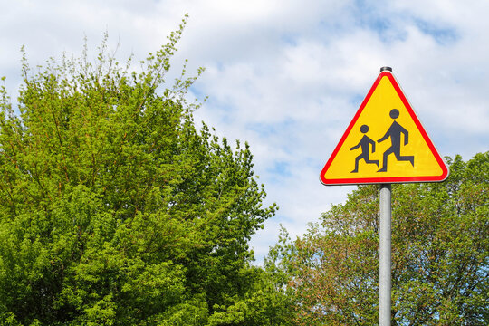 the road sign is made of metal triangular shape in orange color with the image of children against a background of green trees and blue sky with clouds. Careful children